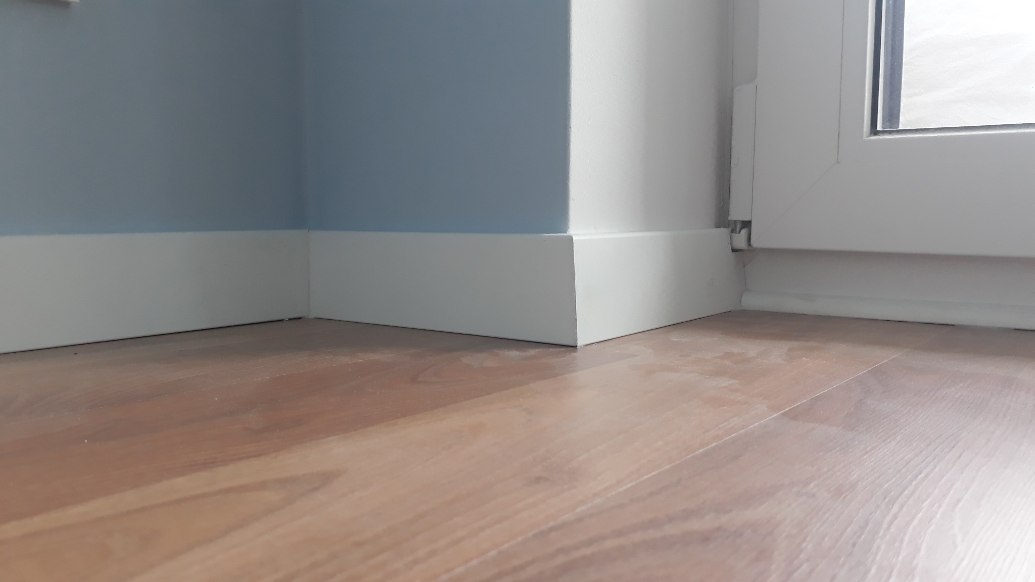 Zamknięty kadr na świeżo położone, białe listwy przypodłogowe w rogu pokoju z panelami podłogowymi i fragmentem okna.