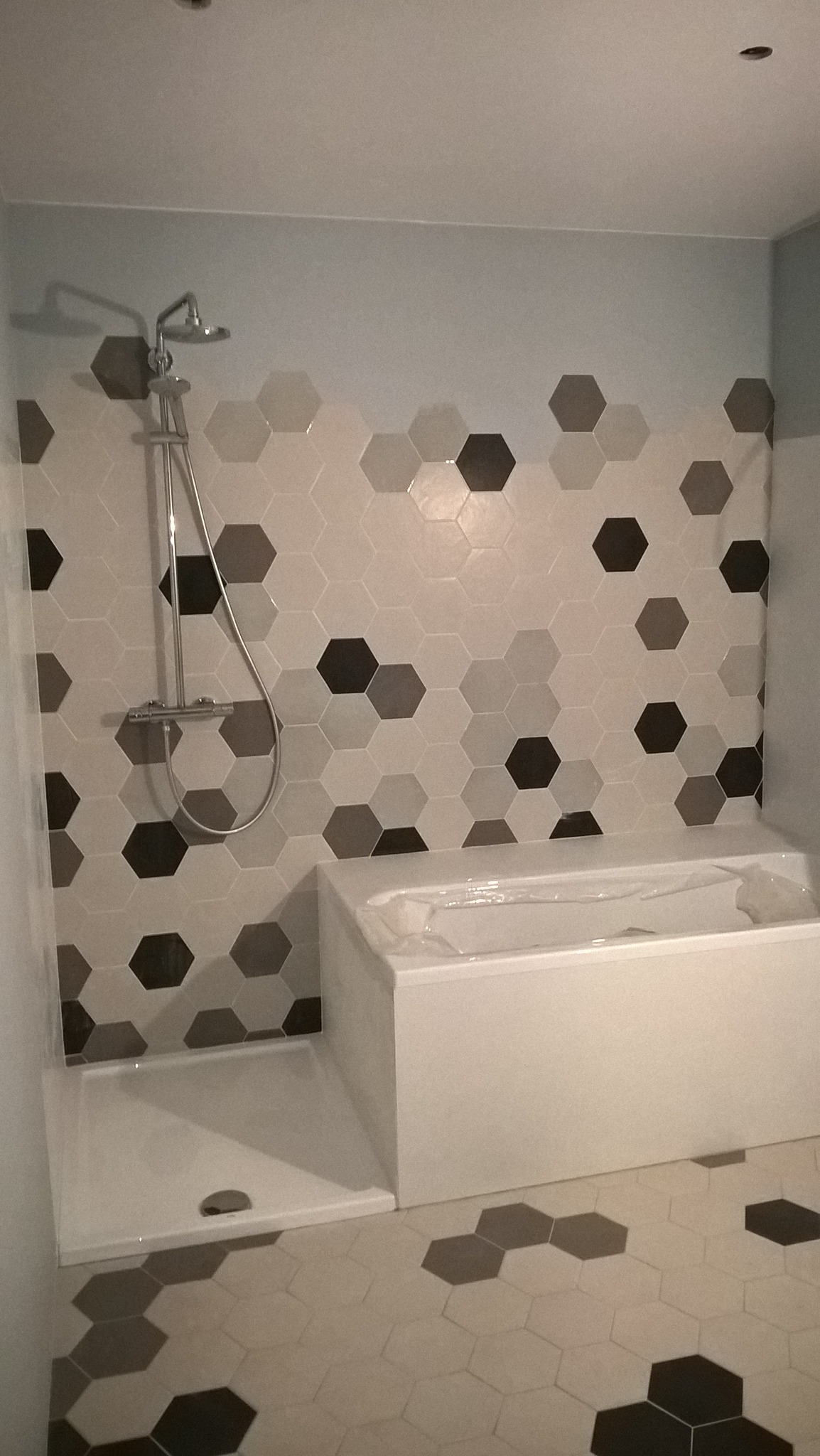 Nowoczesna łazienka z heksagonalnymi płytkami w odcieniach szarości i bieli na ścianach i podłodze, biała wanna zabezpieczona folią ochronną oraz brodzik z odpływem i chromowana bateria prysznicowa.