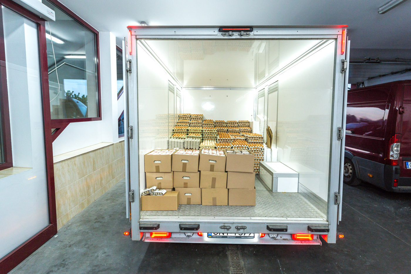 Wnętrze chłodni transportowej wypełnione kartonami i wytłaczankami z jajami. Widoczne światła stop i tablica rejestracyjna pojazdu. Częściowo widoczny drugi samochód.