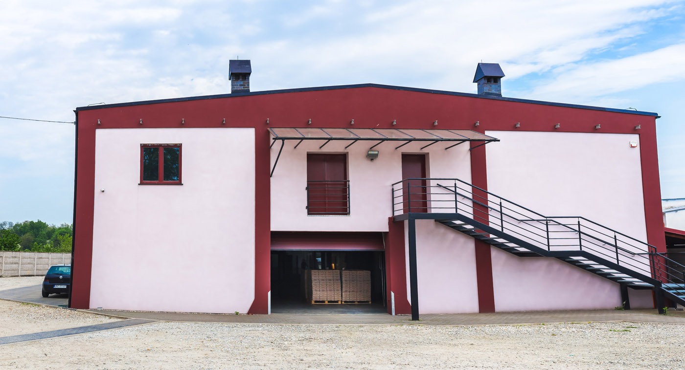 Dwukondygnacyjny budynek magazynowy z rampą załadunkową i schodami zewnętrznymi, pomalowany na biało-bordowo, z paletami wewnątrz. Błękitne niebo w tle.