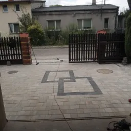 Ułożona kostka brukowa na podjeździe z dekoracyjnym wzorem w kształcie połączonych kwadratów, widoczna otwarta brama wjazdowa z drewnianymi sztachetami i ceglanymi słupkami, narzędzia budowlane w tle.