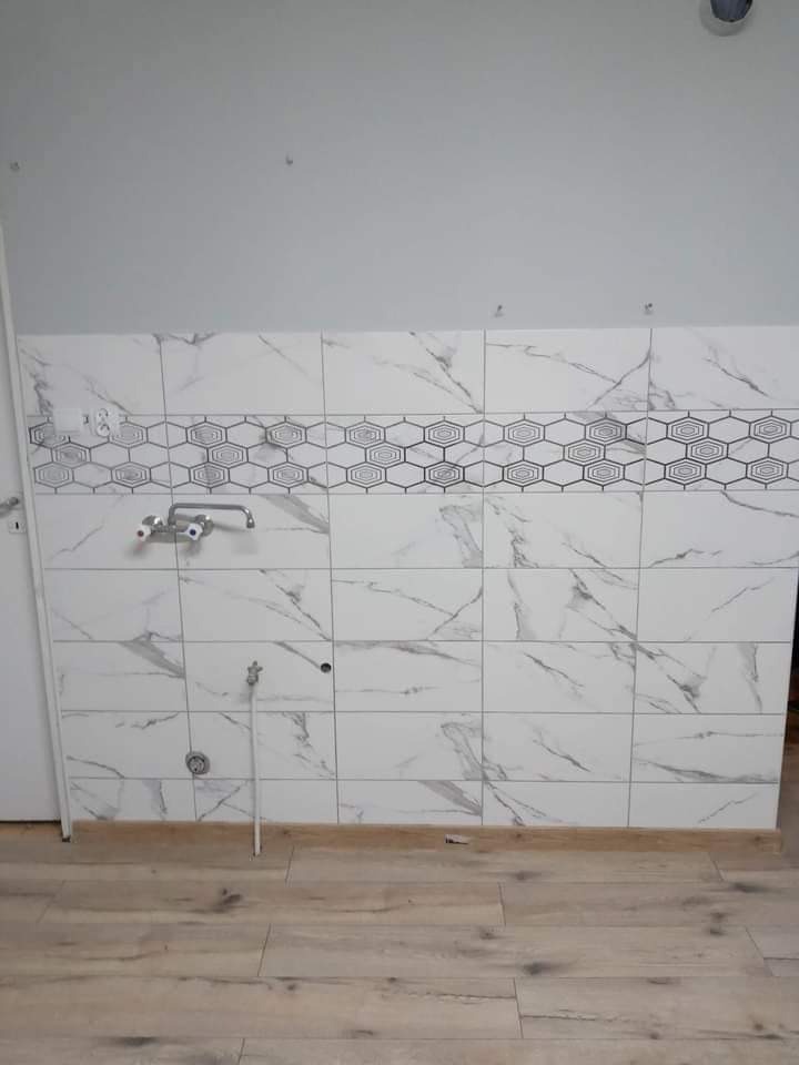 Ściana w łazience wyłożona białymi płytkami z szarym marmurkowym wzorem i pasem dekoracyjnym z motywem heksagonów, widoczna bateria umywalkowa i instalacja wodna przed podłączeniem, podłoga...