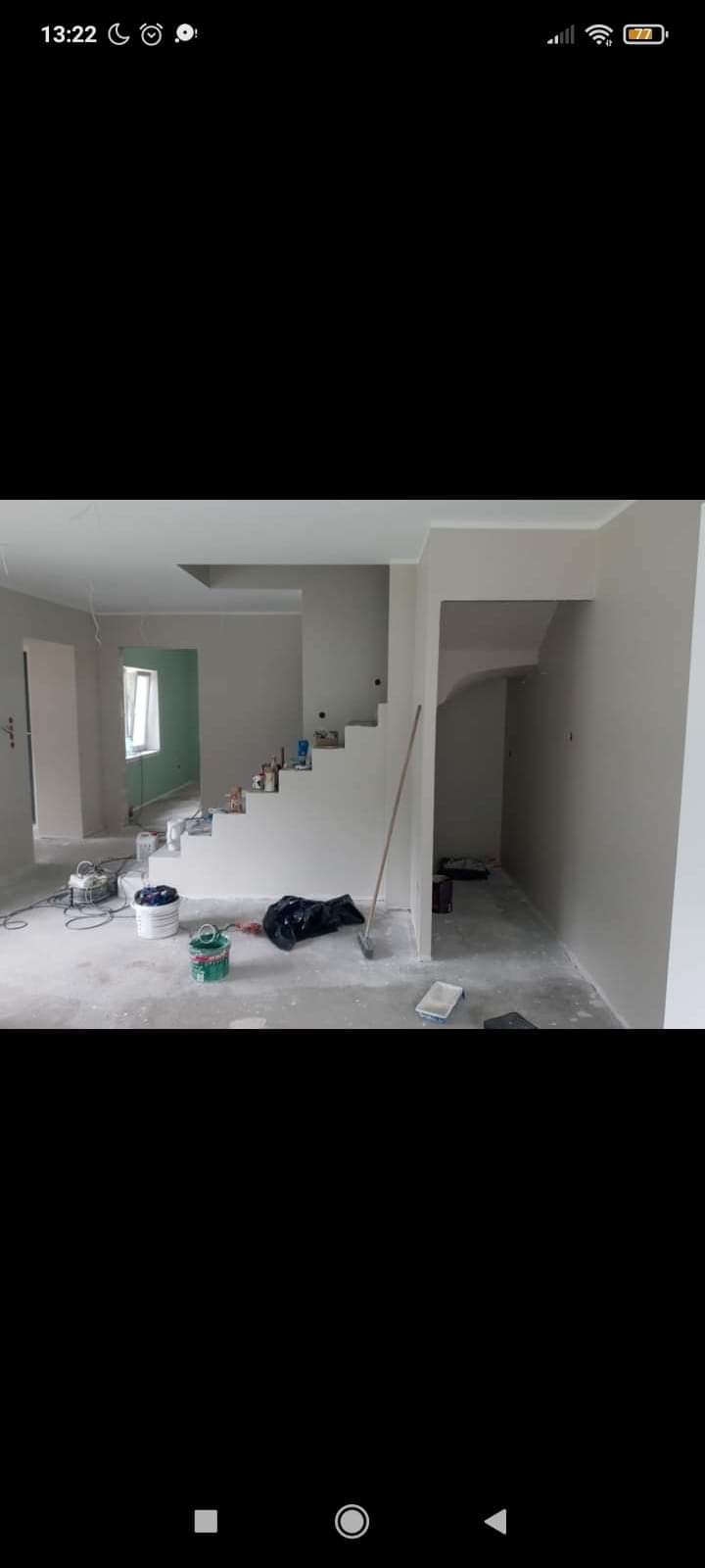 Wnętrze domu w trakcie malowania ścian na szaro, widoczne schody z wiadrami farby, narzędzia malarskie i folia ochronna na podłodze.