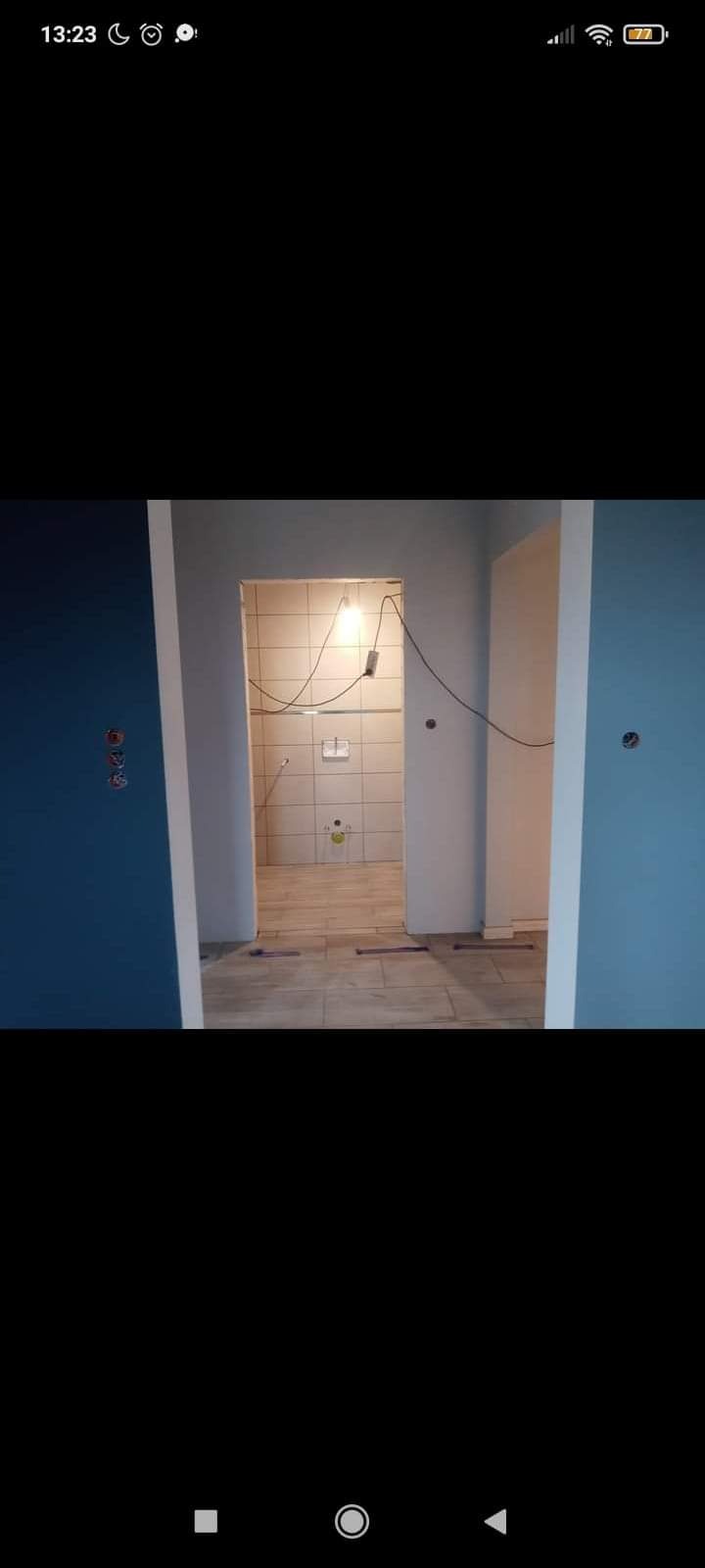 Wnętrze łazienki w trakcie remontu, widoczne płytki na ścianach i podłodze, surowe przewody elektryczne zwisające z sufitu, świeżo pomalowane ściany w kolorach niebieskim i szarym.