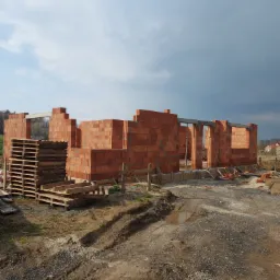 Wznoszone ściany domu z czerwonej ceramiki poryzowanej, widoczne palety z pustakami, przygotowany teren budowy z widocznymi śladami opon i kałużą.