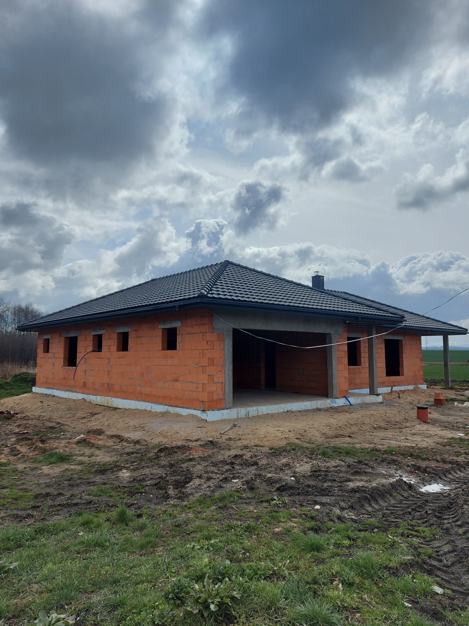 Surowy, jednopiętrowy budynek z czerwonej cegły z ciemną dachówką, w trakcie budowy, z widocznymi otworami okiennymi i niezadaszonym garażem, na tle pochmurnego nieba.