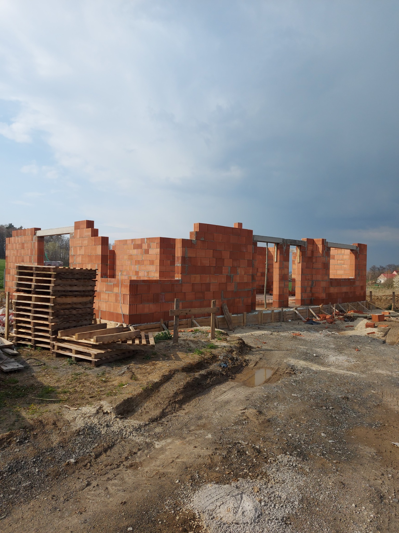 Wznoszone ściany domu z czerwonej ceramiki poryzowanej, widoczne palety z pustakami, przygotowany teren budowy z widocznymi śladami opon i kałużą.