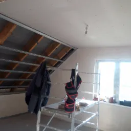 Poddasze w trakcie prac wykończeniowych: widoczne metalowe profile sufitowe, częściowo zamontowane płyty gipsowe, rusztowanie z narzędziami i odzieżą roboczą, okno z jasnym światłem dziennym.