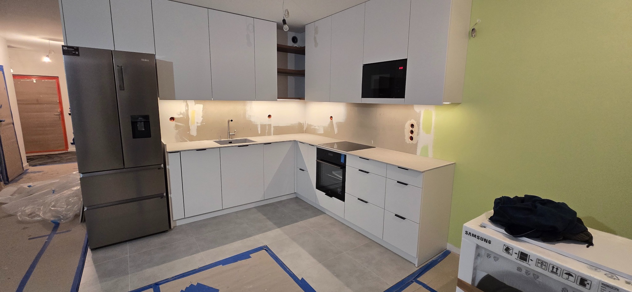 Wnętrze kuchni w trakcie montażu: białe szafki kuchenne, metalowy zlew i płyta indukcyjna, na ścianie widoczne niedokończone gładzie i instalacje elektryczne, obok stoi nowa lodówka i karton...