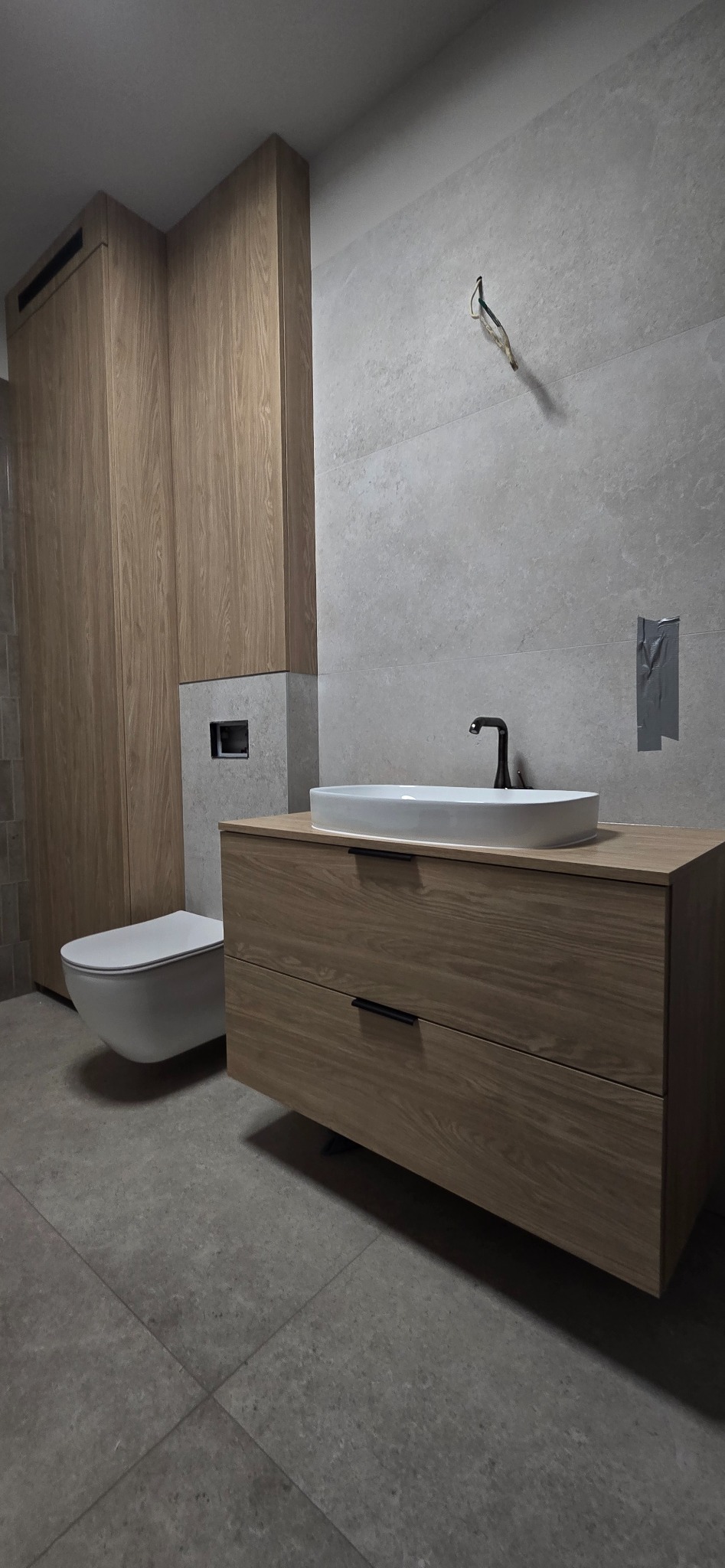 Nowoczesna łazienka z drewnianą szafką pod umywalką, wiszącą toaletą, szarą płytką ścienną i podłogową, widoczne wystające kable elektryczne na ścianie.