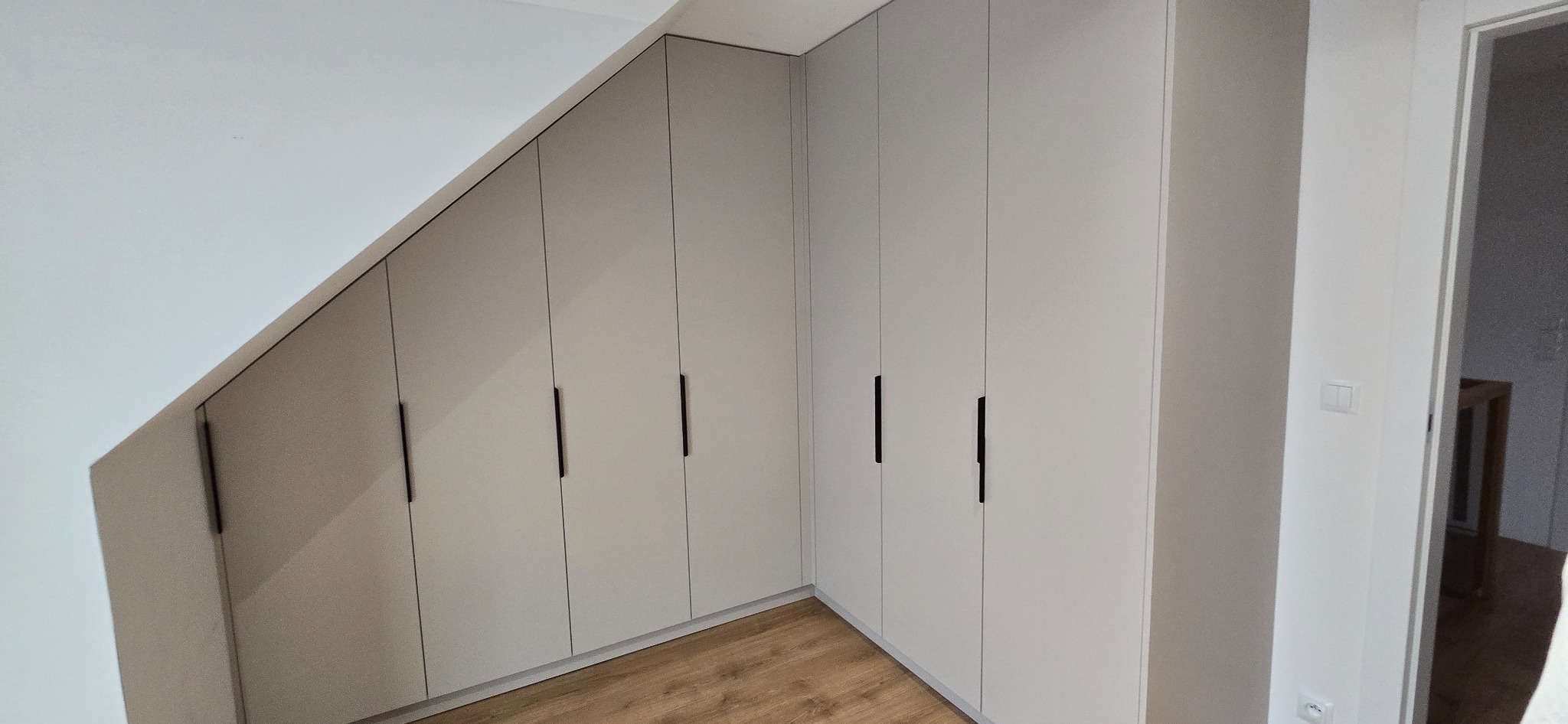 Zabudowa szafy wnękowej na poddaszu ze skośnym sufitem, szare, matowe fronty z czarnymi, minimalistycznymi uchwytami, parkiet na podłodze.