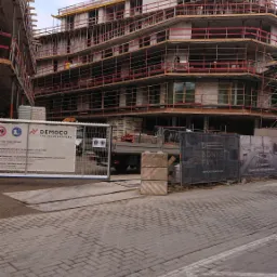 Budowa wielopiętrowego budynku mieszkalnego z widocznym rusztowaniem, dźwigiem i tablicami informacyjnymi na pierwszym planie.