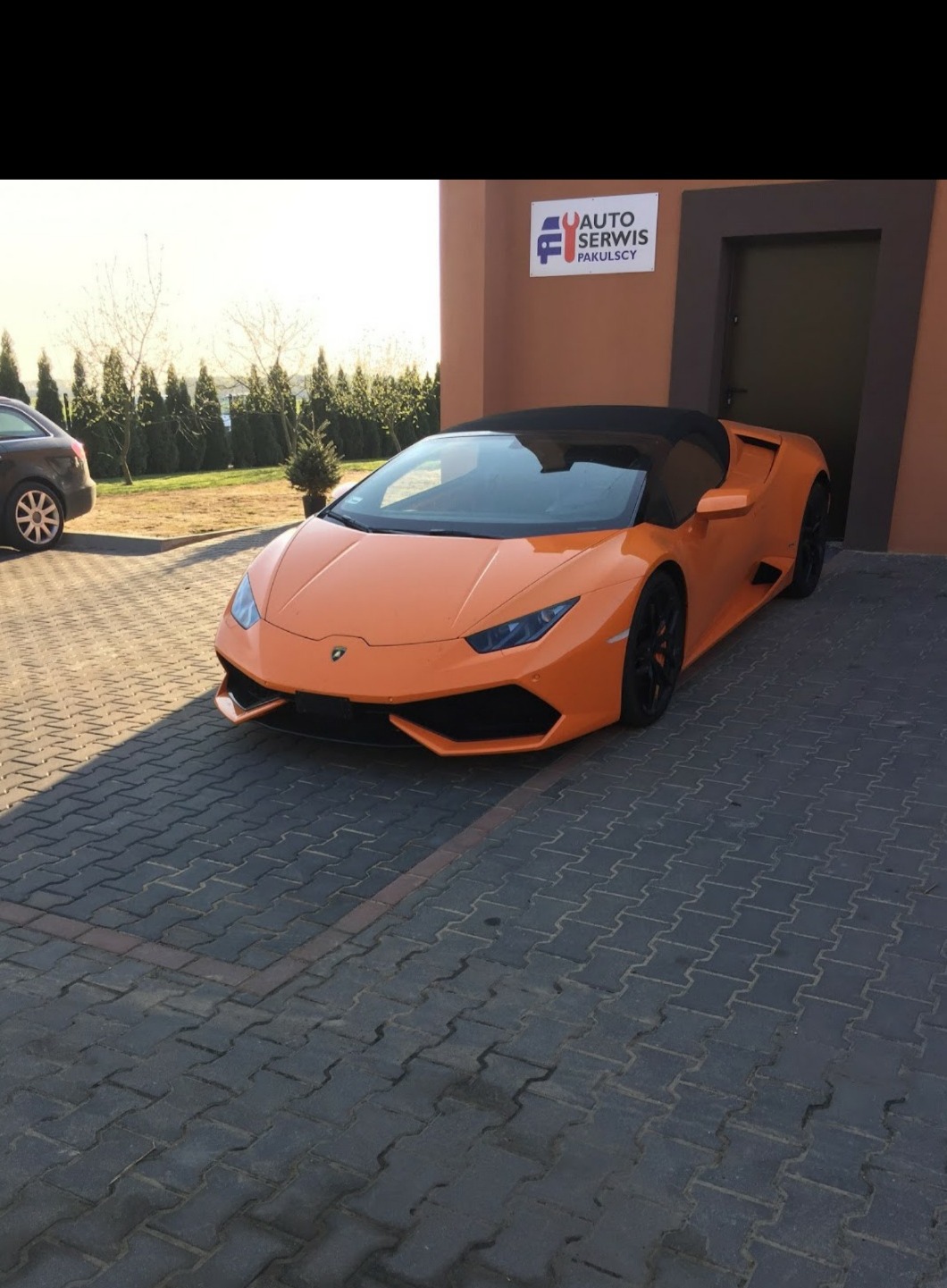 Pomarańczowe Lamborghini Huracan Spider zaparkowane przed budynkiem warsztatu samochodowego z logo Auto Serwis Pakulscy.
