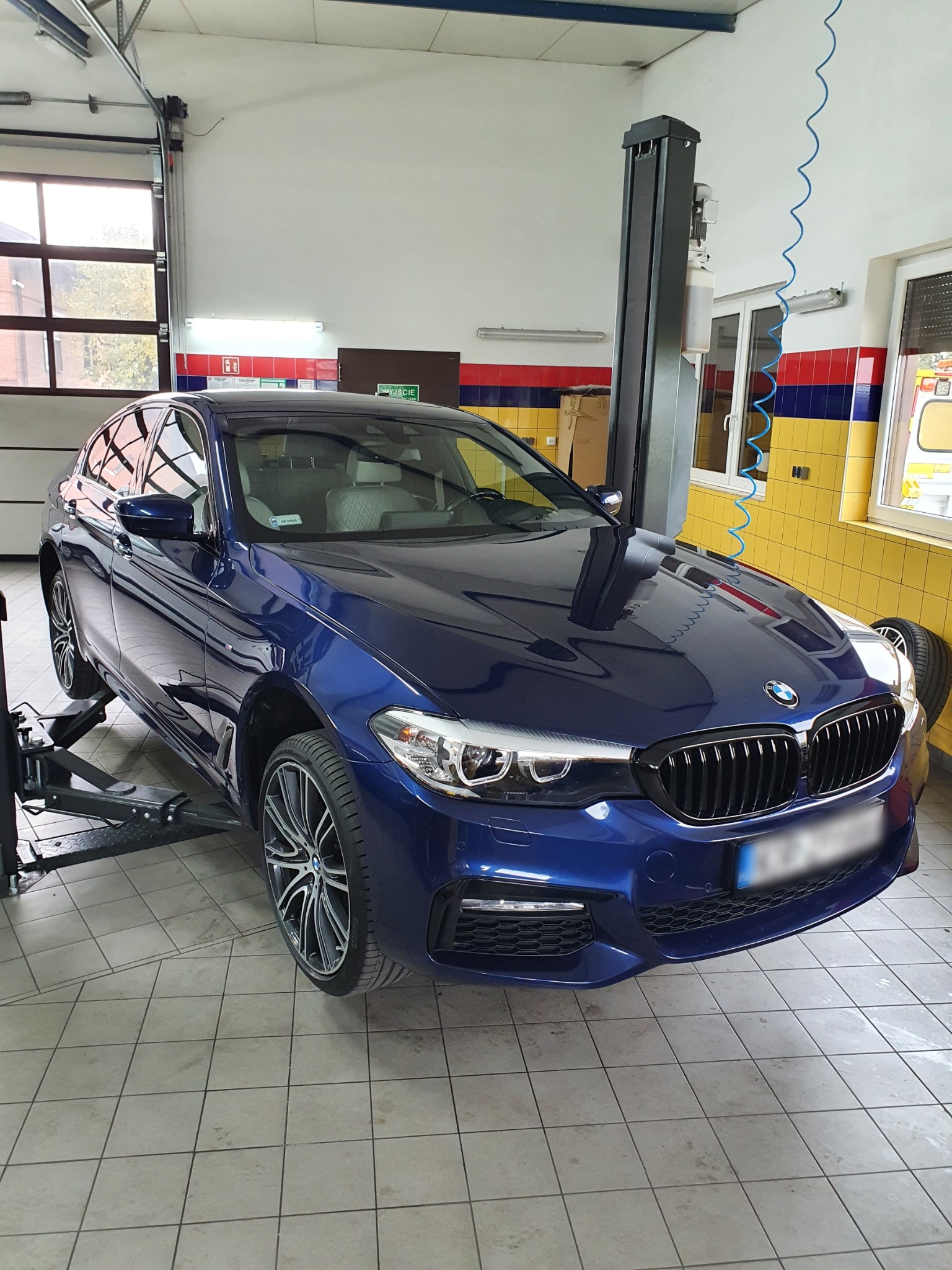 Niebieski samochód BMW serii 5 na podnośniku w warsztacie samochodowym, widoczna opona z boku, płytki na podłodze i narzędzia w tle.