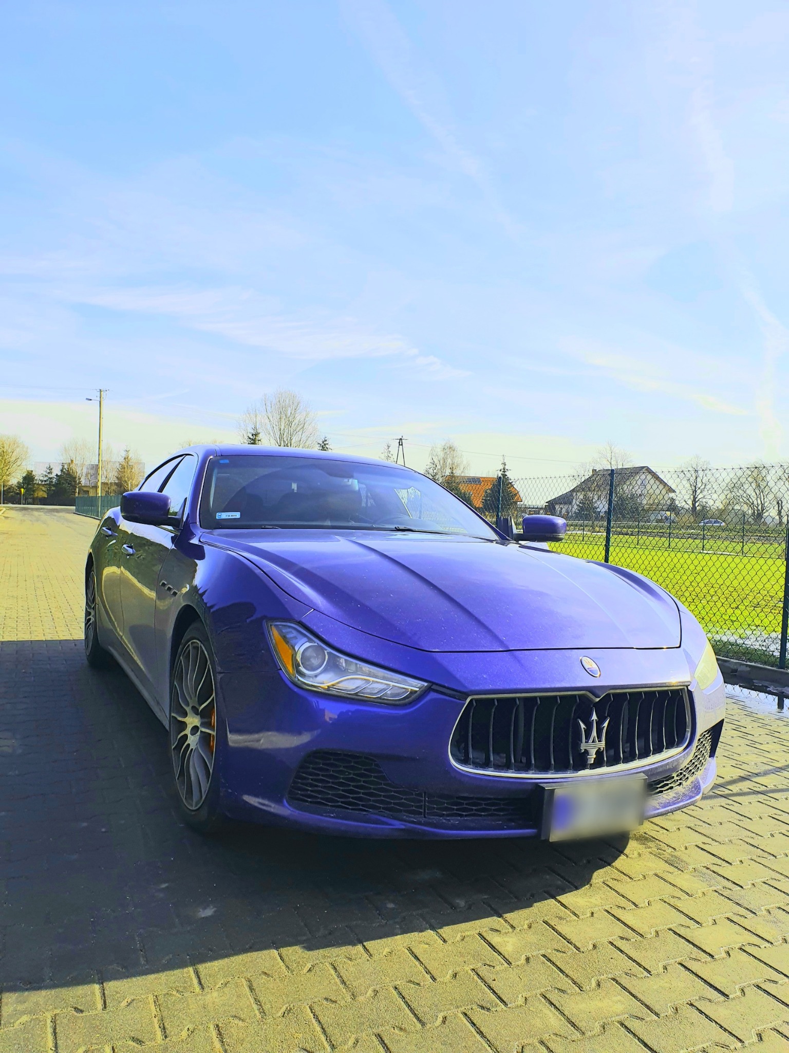 Granatowy samochód sportowy marki Maserati zaparkowany na brukowanej drodze w słoneczny dzień, widok z przodu z lekko uniesionym kątem, z widocznymi detalami grilla i logo.