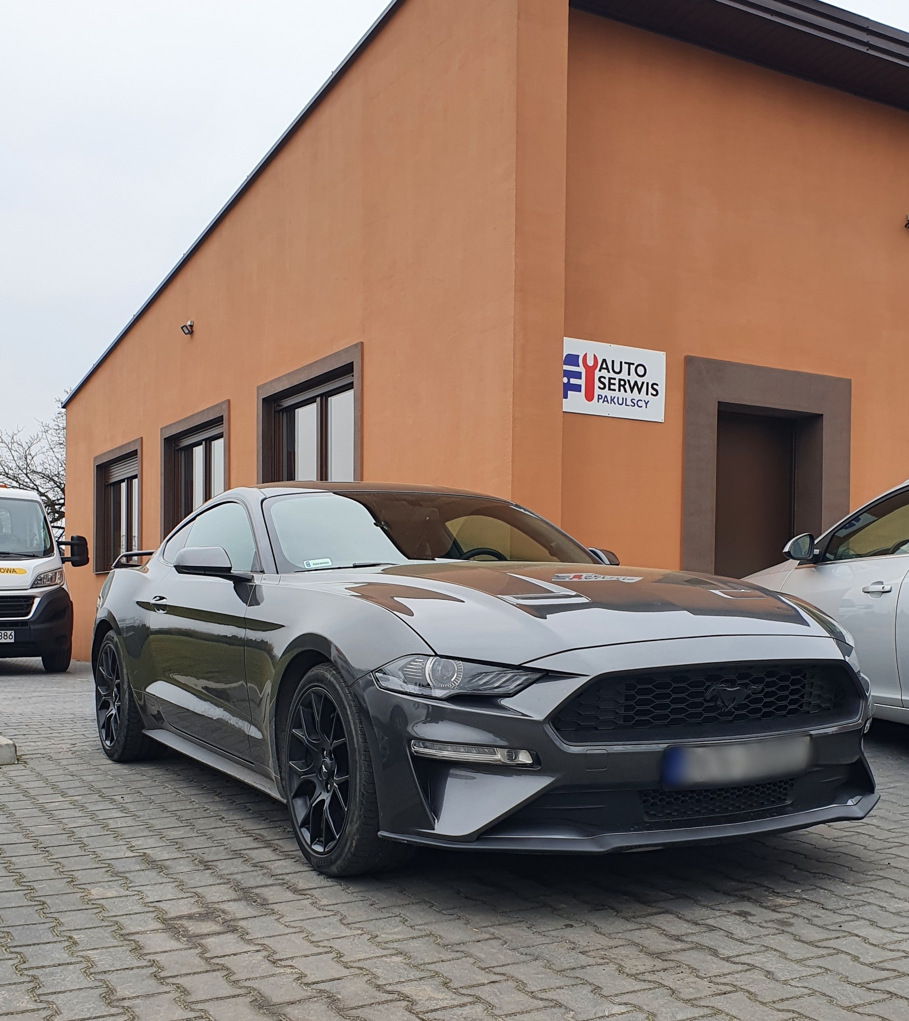 Grafitowy Ford Mustang z czarnymi felgami stoi przed budynkiem auto serwisu z pomarańczową elewacją i logo firmy.