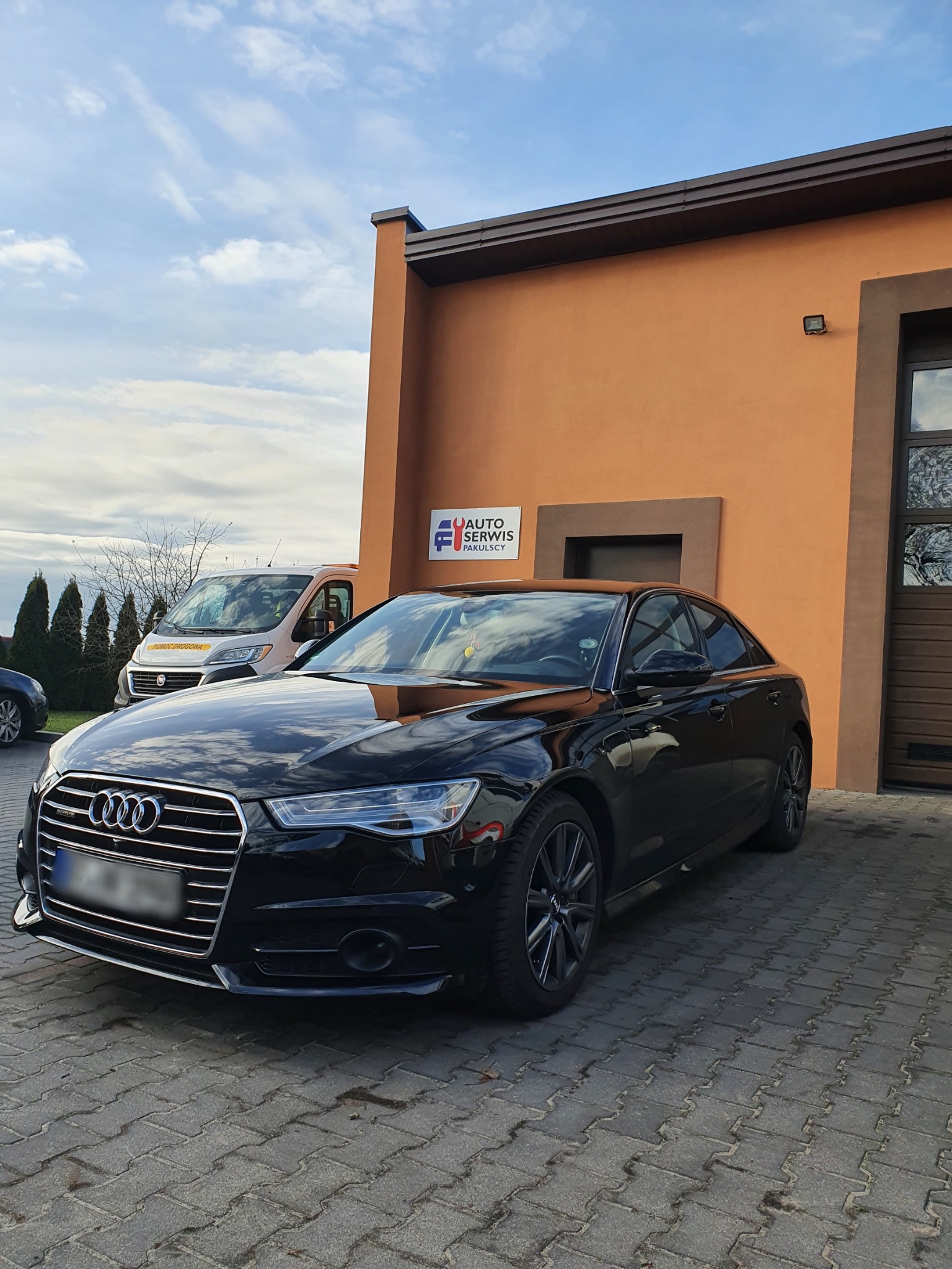 Czarny samochód Audi A6 zaparkowany przed budynkiem serwisu samochodowego z pomarańczową elewacją i logo. W tle widoczny bus pomocy drogowej.