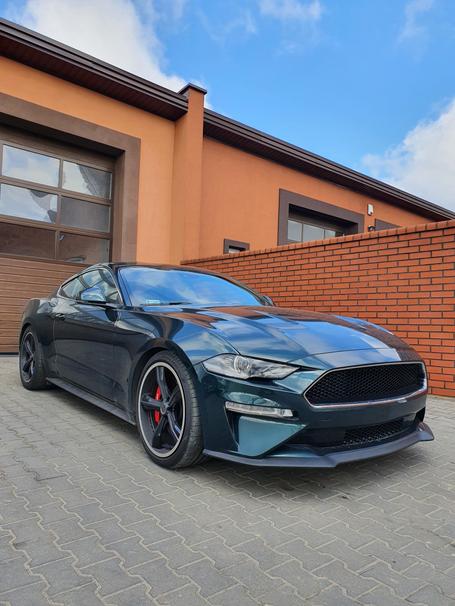 Ciemnozielony Ford Mustang z białym pasem na masce, zaparkowany na kostce brukowej przed budynkiem z ceglaną ścianą i garażem, widoczne czerwone zaciski hamulcowe.