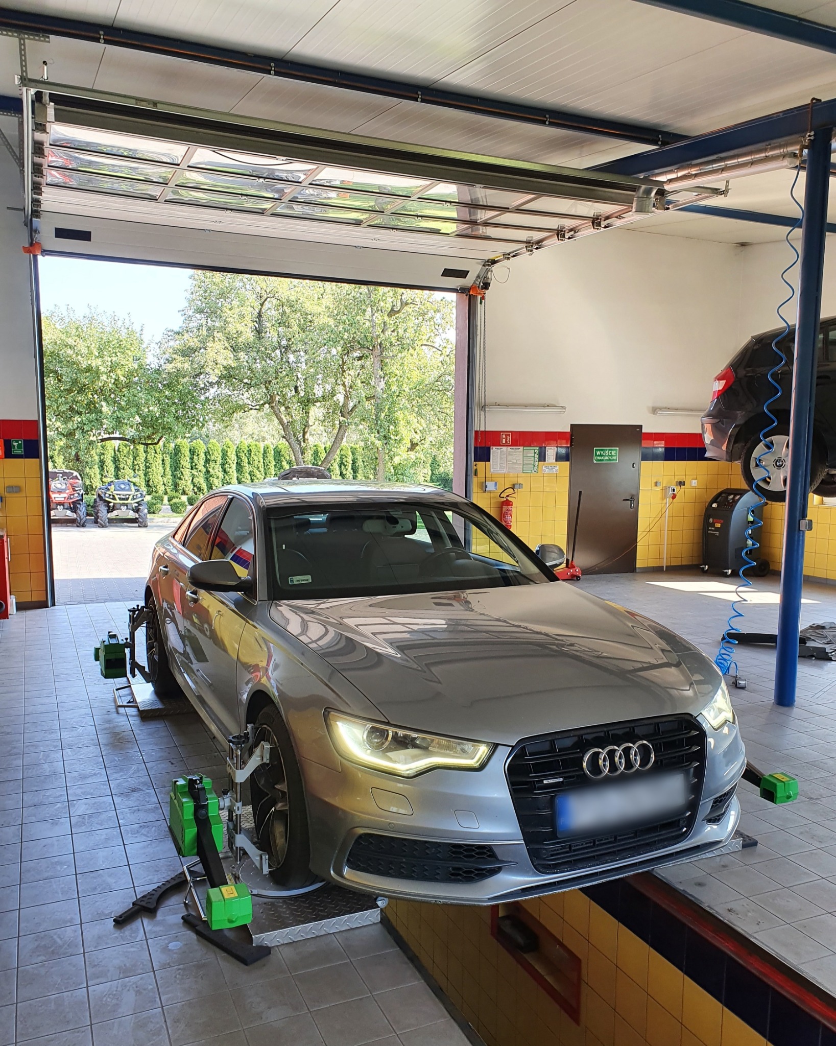 Srebrny samochód Audi A6 na stanowisku do geometrii kół w warsztacie, z widocznymi czujnikami i urządzeniami pomiarowymi, otwartą bramą garażową i pojazdami terenowymi w tle.