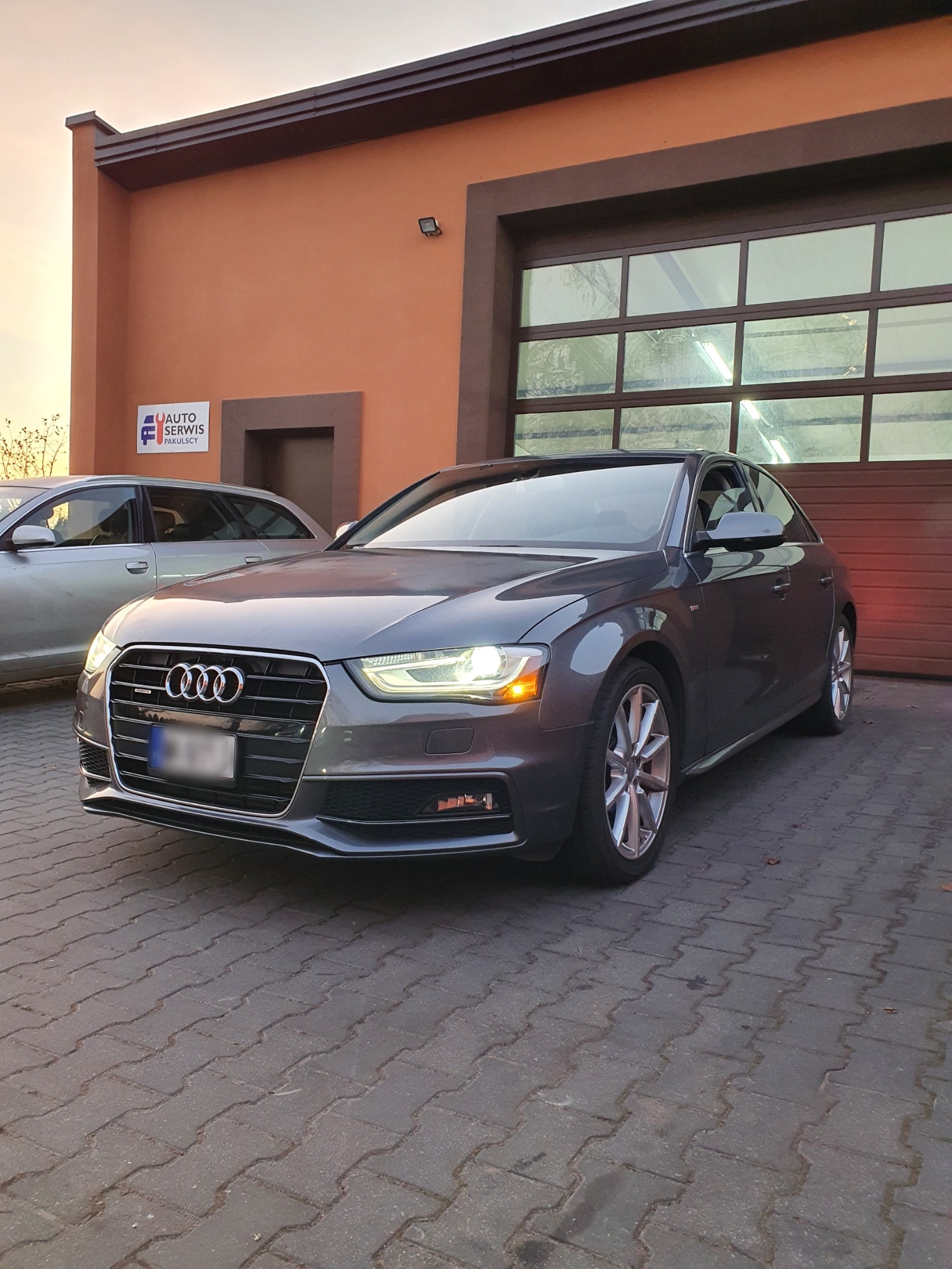 Szary samochód Audi A4 zaparkowany przed budynkiem warsztatu z przeszklonymi drzwiami garażowymi i szyldem 'Auto Serwis Pakulscy'.