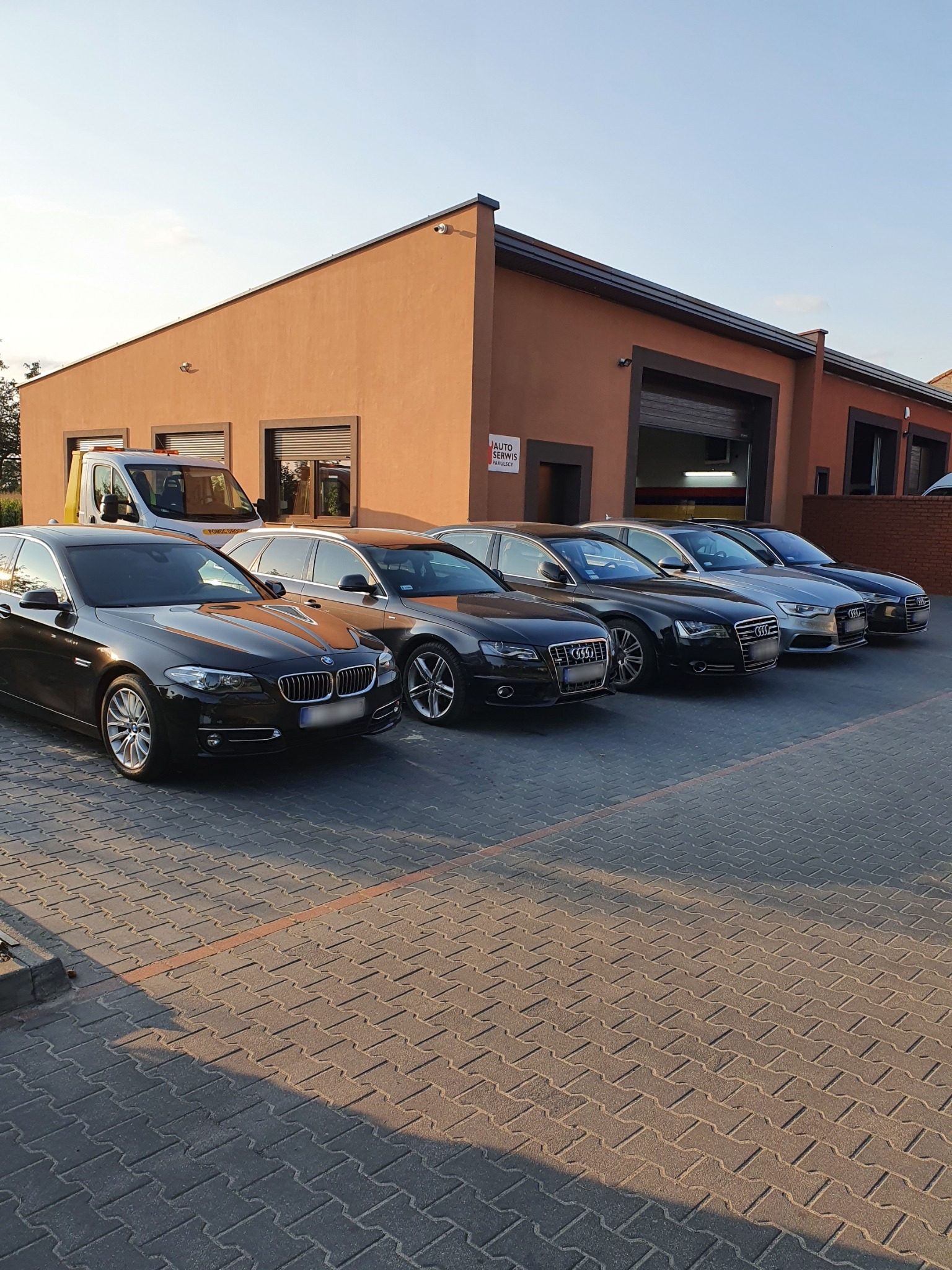 Rząd zaparkowanych samochodów marki Audi i BMW przed budynkiem serwisu samochodowego w ciepłym świetle zachodzącego słońca; widoczny także bus serwisu z napisem 'Pomoc Drogowa'.