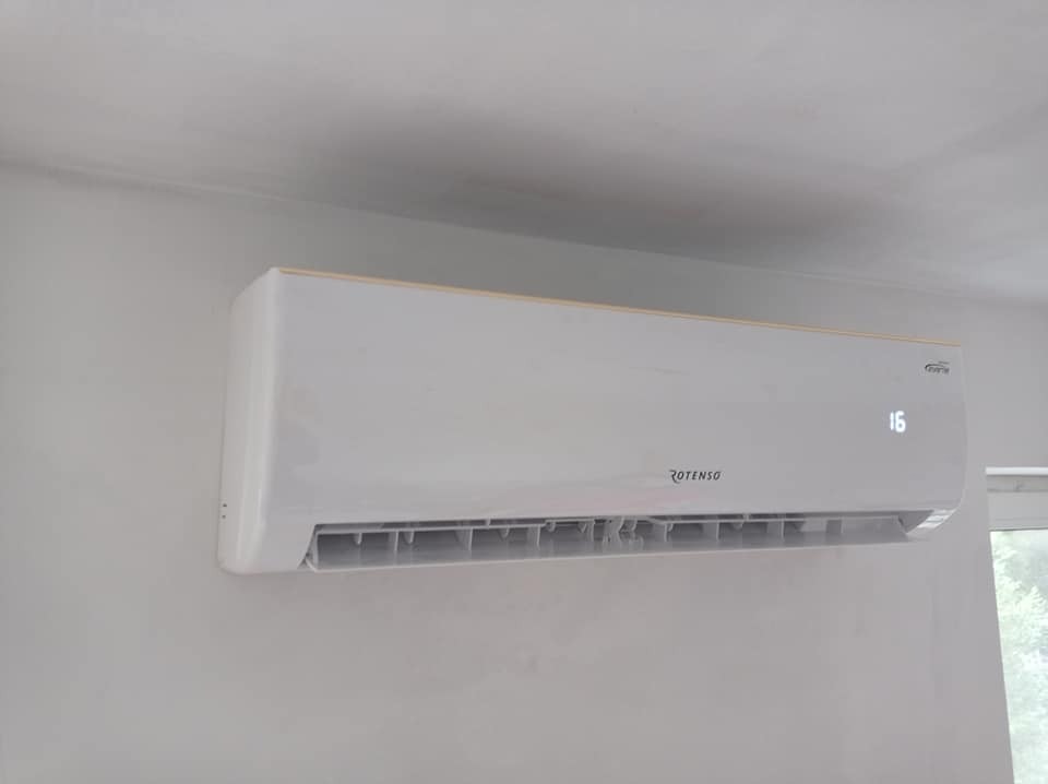 Biała klimatyzacja ścienna Rotenso z wyświetlaczem temperatury 16 stopni, zamontowana na szarej ścianie w pomieszczeniu.