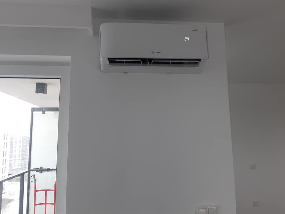 Biały klimatyzator ścienny z wyświetlaczem temperatury 24 stopnie Celsjusza, zamontowany w jasnym, nowoczesnym wnętrzu obok przeszklonych drzwi balkonowych.