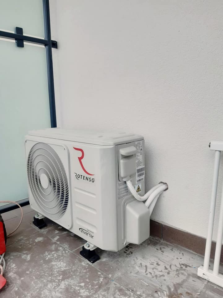 Biała jednostka zewnętrzna klimatyzatora Rotenso Inverter, zamontowana na czarnych podstawkach, z przewodami wchodzącymi w ścianę. Widoczne ślady wiercenia w podłodze.