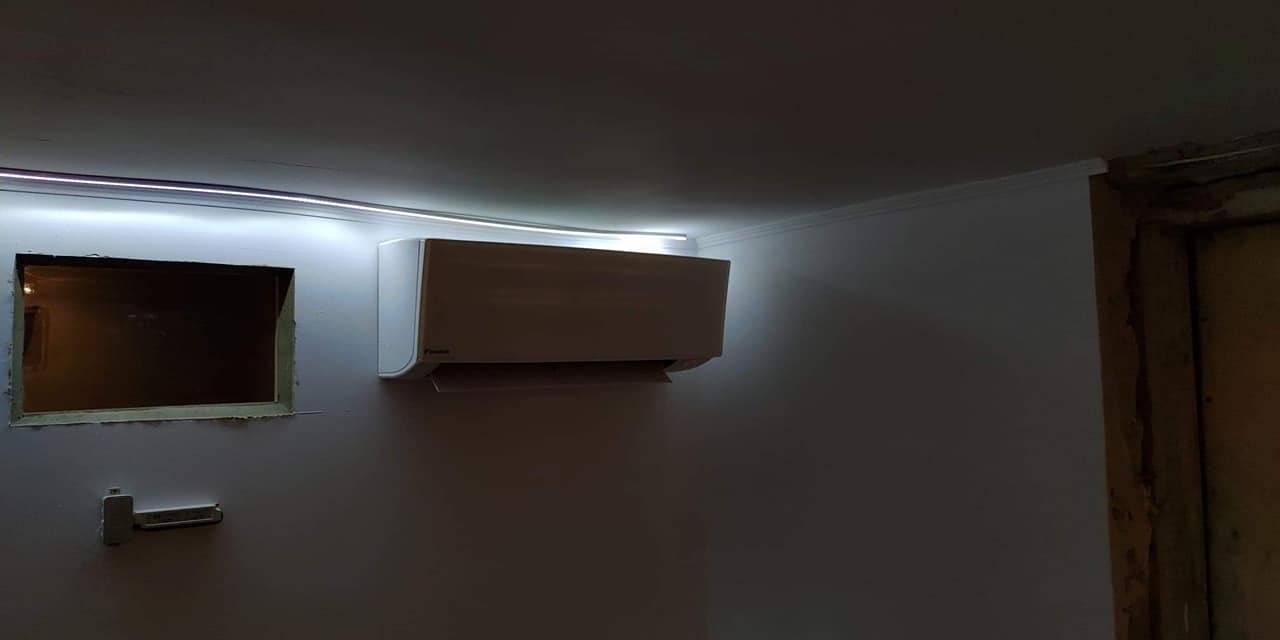 Biały klimatyzator ścienny zamontowany w pokoju z surowym oknem i listwą LED, widoczne ślady prac wykończeniowych.