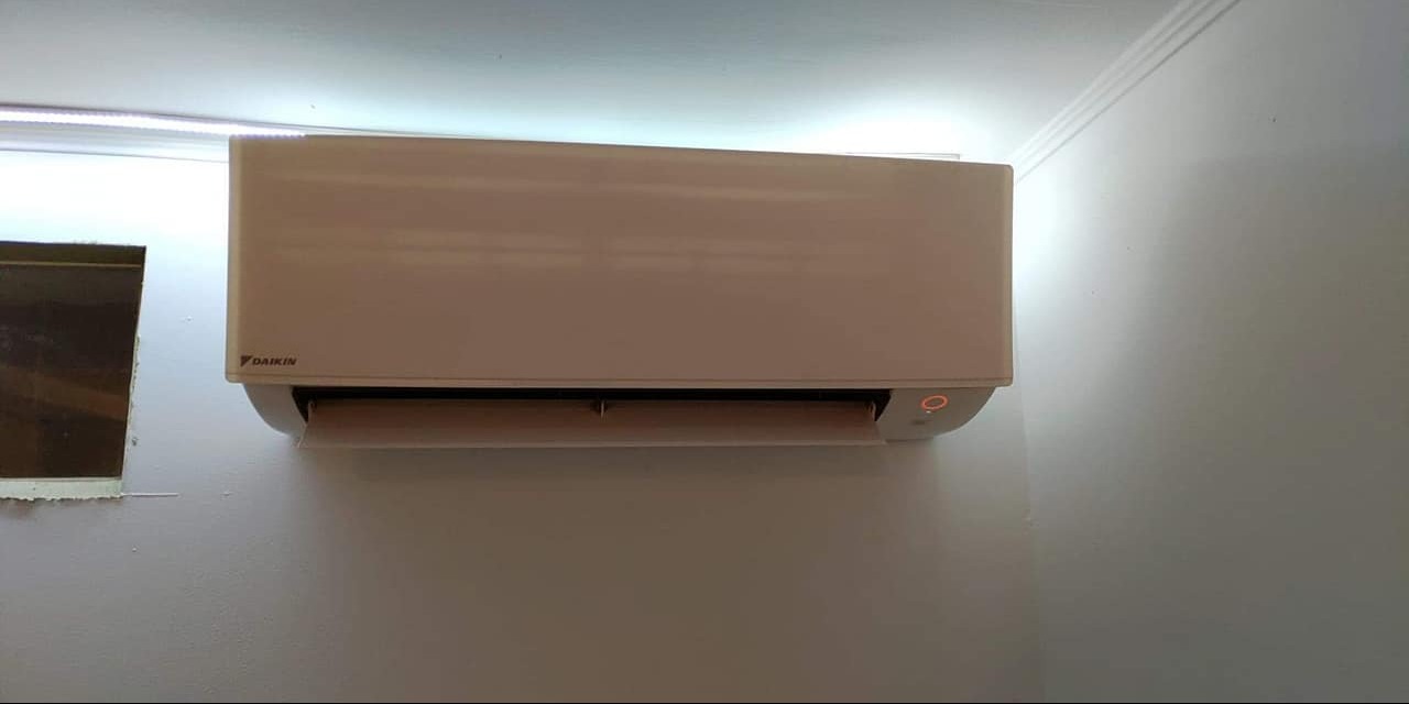 Biała jednostka klimatyzacji Daikin zamontowana na ścianie w pomieszczeniu, widoczna dioda sygnalizacyjna i fragment okna z parapetem w tle.