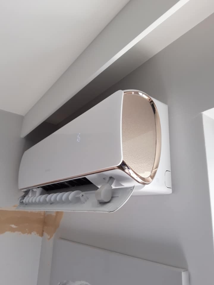 Biały klimatyzator ścienny z elementami w kolorze różowego złota, zamontowany na szarej ścianie, z widocznymi śladami po niedokończonym malowaniu.