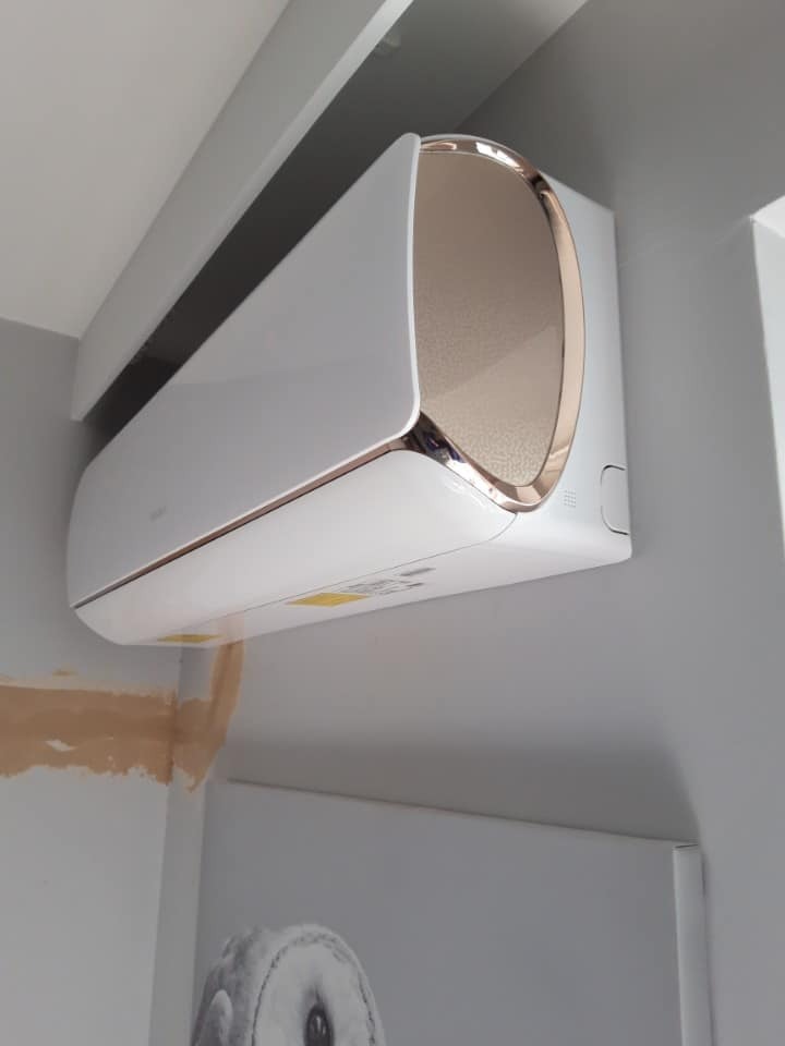 Biała klimatyzacja z elementami w kolorze złotym, zamontowana na szarej ścianie w pomieszczeniu z widocznym fragmentem obrazu z motywem sowy.