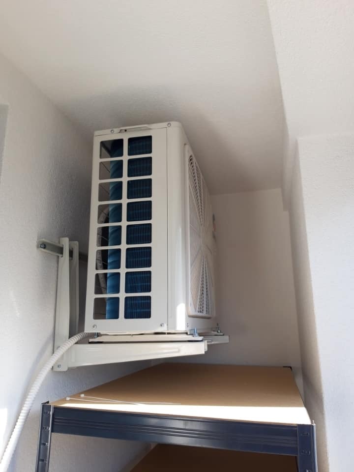 Klimatyzator ścienny zamontowany na wspornikach w pomieszczeniu w Warszawie, widoczna jednostka zewnętrzna i fragment regału pod spodem.