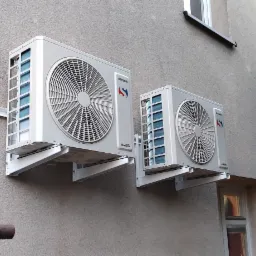 Dwa klimatyzatory Sinclair zamontowane na szarej, teksturowanej ścianie budynku z widocznymi oknami i rynną.