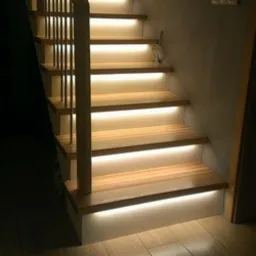 Drewniane schody z podświetleniem LED zamontowanym pod każdym stopniem, tworzącym ciepłą, rozproszoną iluminację. Widoczne pionowe barierki i fragment ściany w tle.