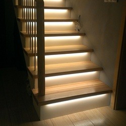 Drewniane schody z podświetleniem LED zamontowanym pod każdym stopniem, tworzącym ciepłą, rozproszoną iluminację. Widoczne pionowe barierki i fragment ściany w tle.