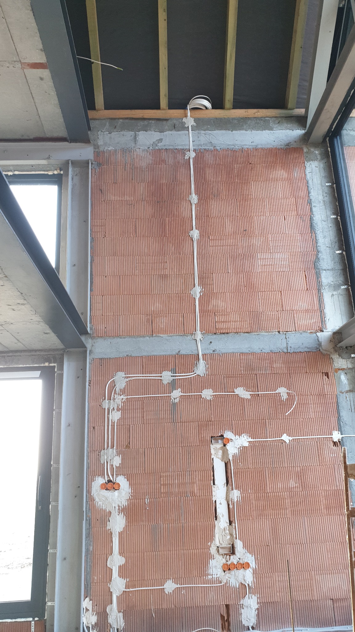 Surowa ściana z pustaków ceramicznych w trakcie montażu przewodów elektrycznych, widoczne puszki instalacyjne zalane gipsem i mocowania kabli, w tle stalowa konstrukcja szkieletowa budynku.