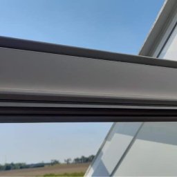 Przemysłowy Serwis Sprzątający - Fragment okna dachowego marki OKPOL z widoczną ramą i szybą, na tle błękitnego nieba i zieleni drzew.