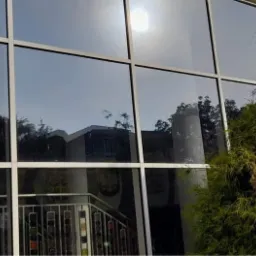 Duże, panoramiczne okno z białymi ramami odbija budynek i niebo; z zewnątrz widoczne krzewy i iglak.