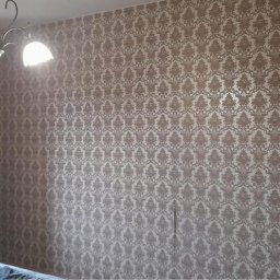 Przemysłowy Serwis Sprzątający - Ściana pokryta beżową tapetą w ozdobny wzór, widoczny fragment okna z zasłoną w pasy i dekoracyjny piec kaflowy w odcieniach zieleni.