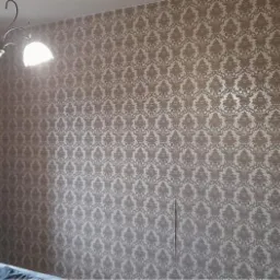 Ściana pokryta beżową tapetą w ozdobny wzór, widoczny fragment okna z zasłoną w pasy i dekoracyjny piec kaflowy w odcieniach zieleni.