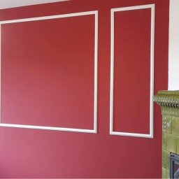 Przemysłowy Serwis Sprzątający - Czerwona ściana z białymi, dekoracyjnymi listwami, odkurzacz w rogu, widoczny fragment pieca kaflowego i lampa sufitowa