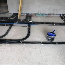 Szare, surowe wnętrze w trakcie prac wykończeniowych. Na podłodze ułożone czarne kable elektryczne, przymocowane białymi opaskami. W tle drabina oparta o ścianę. Wiaderko z niebieskim pędzlem po...