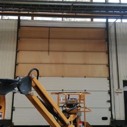 Przemysłowy Serwis Sprzątający - Wnętrze hali przemysłowej z bramą segmentową, częściowo wyczyszczoną z brudu, widoczną z podnośnika koszowego.