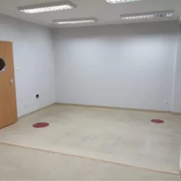 Pusta, jasna sala z beżową podłogą i białymi ścianami, z drzwiami z okrągłym okienkiem po lewej stronie i dwoma czerwonymi okręgami na podłodze.