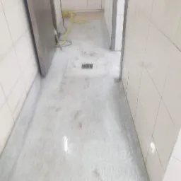 Wnętrze pomieszczenia z białymi kafelkami, mokra posadzka z widocznym odpływem liniowym, żółty przewód na podłodze.