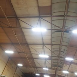 Przemysłowy Serwis Sprzątający - Wnętrze hali przemysłowej z metalową konstrukcją dachu, oświetlone podłużnymi lampami LED, widoczne fragmenty ścian z panelami.