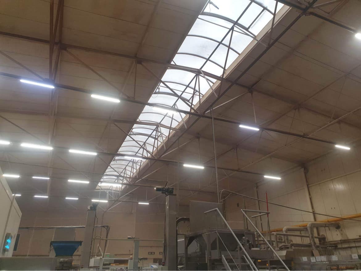 Wnętrze hali produkcyjnej z widocznym oświetleniem LED, konstrukcją dachu z świetlikami i elementami linii produkcyjnej.