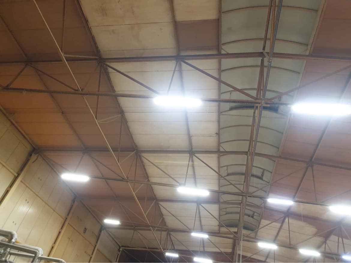 Wnętrze hali przemysłowej z metalową konstrukcją dachu, oświetlone podłużnymi lampami LED, widoczne fragmenty ścian z panelami.