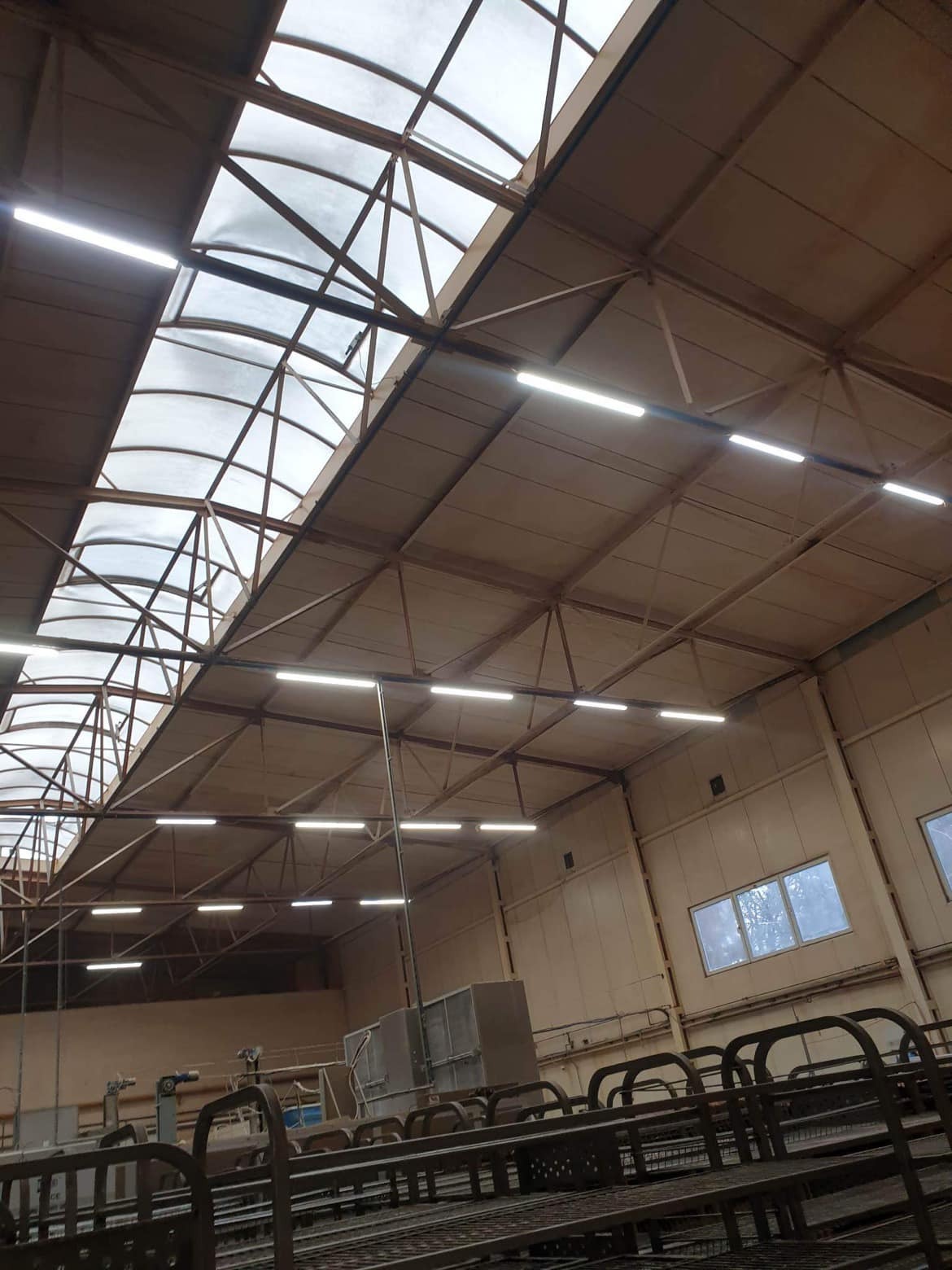 Wnętrze hali przemysłowej z widoczną konstrukcją dachu z świetlikami i szeregiem lamp LED, metalowe konstrukcje na pierwszym planie.